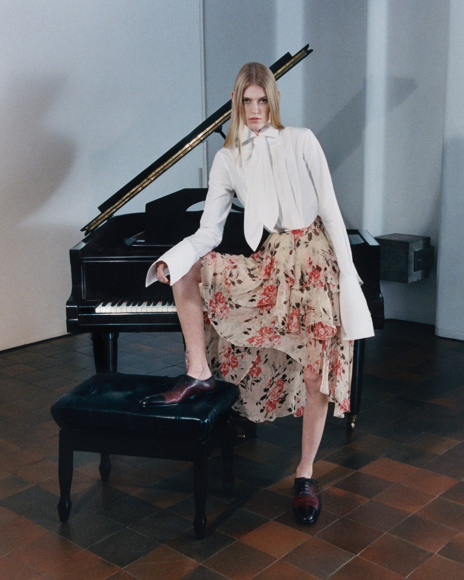 Loewe SS25 Online Stories Blond woman wearing a white blouse and a floral skirt in front of a piano